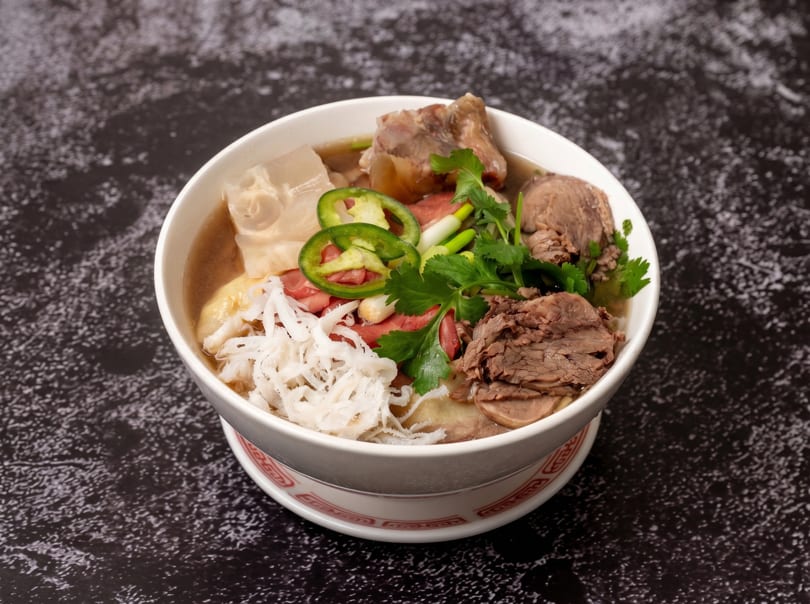 A bowl of Vietnamese pho with beef, tripe, cilantro, and sliced green chilies on a dark surface.