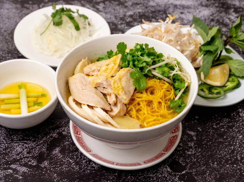 A bowl of chicken noodle soup with sides of bean sprouts, herbs, and dipping sauce.