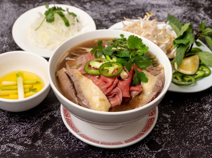 A bowl of pho with beef, cilantro, and jalapeños, surrounded by side dishes.