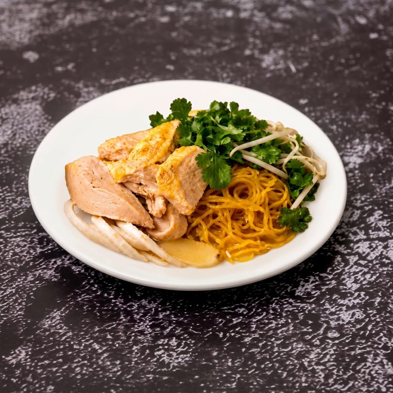 A white plate holds chicken slices, yellow noodles, cilantro, and bean sprouts on a dark textured surface.